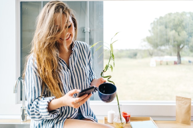 Mooie jonge vrouw die op het aanrecht zit en op de mobiele telefoon bladert in de moderne keuken