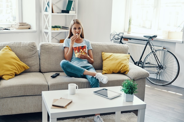 Mooie jonge vrouw die popcorn eet terwijl ze thuis tv kijkt op de bank