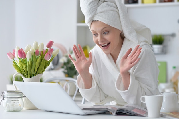 Mooie jonge vrouw die thuis een witte badjas met laptop draagt