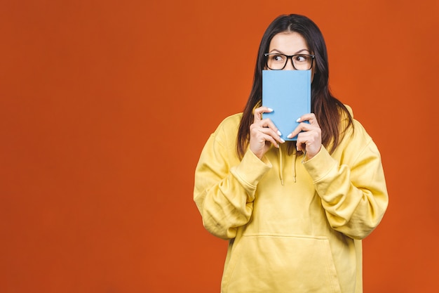 Mooie jonge vrouw die toevallige status draagt die over oranje achtergrond wordt geïsoleerd, die een boek leest.