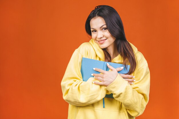 Mooie jonge vrouw die toevallige status draagt die over oranje achtergrond wordt geïsoleerd, die een boek leest.