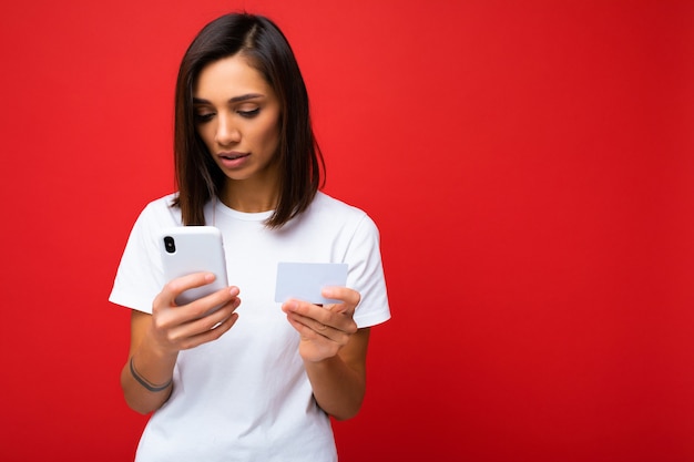 Mooie jonge vrouw die vrijetijdskleding draagt die geïsoleerd over achtergrond staat die op internet surft via de telefoon die naar het mobiele scherm kijkt