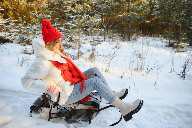 Mooie jonge vrouw die warme winterkleren draagt, op een slee zit en van de heuvel glijdt, plezier heeft op een besneeuwde winterdag