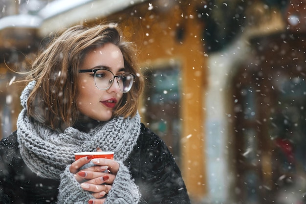 Mooie jonge vrouw draagt gebreide sjaal en bril wandelen in de stad tijdens sneeuwval. Lege ruimte