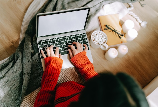 Mooie jonge vrouw draagt gebreide warme trui zittend op de vloer en werkt of leert online op internet op laptop met kop warme drank thuis Gezellige winter versierd met kaarsen Bovenaanzicht