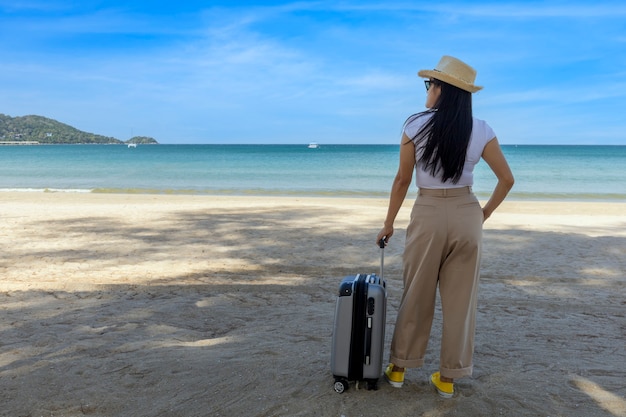 Mooie jonge vrouw draagt t-shirt, lange broek en strooien hoed met een koffer op een tropisch strand.