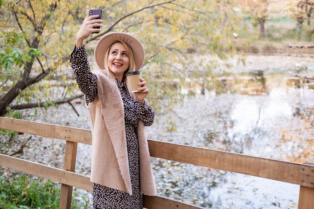 Mooie jonge vrouw drinkt koffie en neemt selfie op telefoon in de natuur in het herfstpark in de herfst