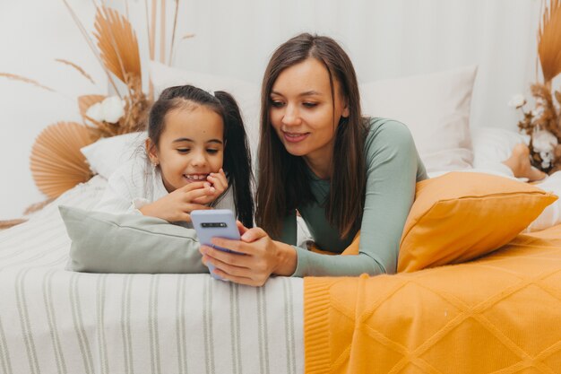 Mooie jonge vrouw en haar dochtertje kijken naar de telefoon terwijl ze thuis in bed liggen