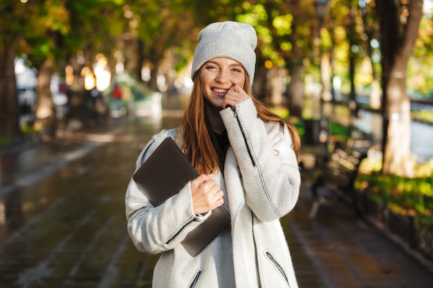 Mooie jonge vrouw gekleed in herfst jas en hoed buiten wandelen, met laptopcomputer