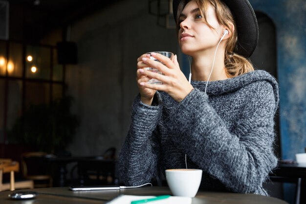 Mooie jonge vrouw gekleed in trui en hoed zit aan de café tafel binnenshuis, luisteren naar muziek met koptelefoon, koffie drinken