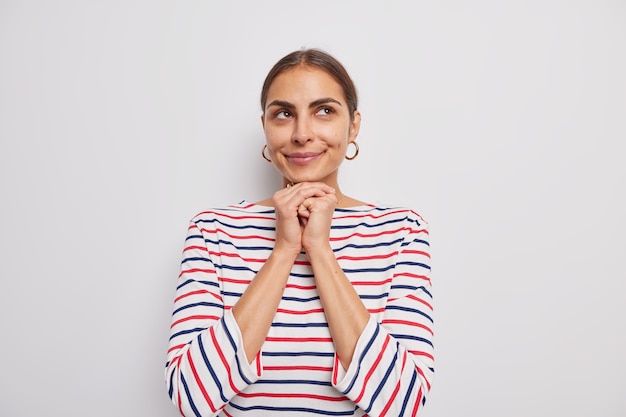 Mooie jonge vrouw houdt handen onder kin glimlacht zachtjes kijkt boven kijkt met bewondering draagt casual gestreepte trui geïsoleerd over witte muur denkt aan iets moois