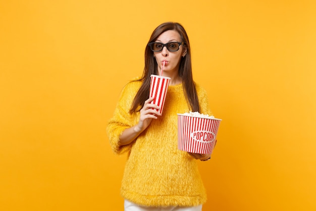 Mooie jonge vrouw in 3d imax-bril kijken naar filmfilm met emmer popcorn, cola of frisdrank drinken uit plastic beker geïsoleerd op gele achtergrond. Mensen oprechte emoties in de bioscoop, levensstijl.