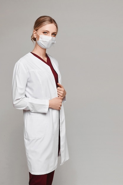 Mooie jonge vrouw in beschermend masker en het medische eenvormige stellen op grijze geïsoleerde achtergrond ,. Vrouwelijke arts in witte medische jas. Gezondheidszorg concept