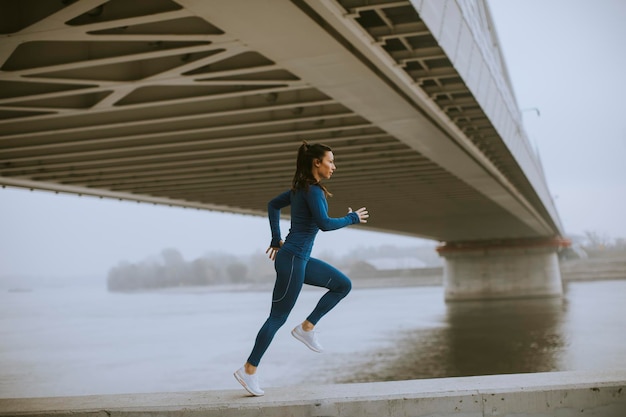 Mooie jonge vrouw in blauw trainingspak die op herfstochtend langs de rivier loopt