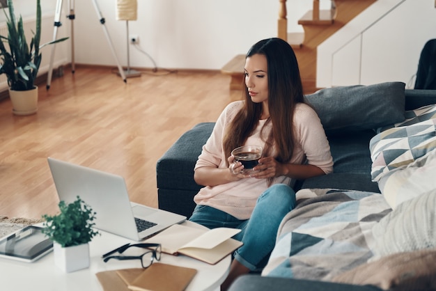 Mooie jonge vrouw in casual kleding die laptop gebruikt terwijl ze thuis tijd doorbrengt