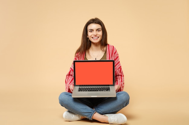 Mooie jonge vrouw in casual kleding zitten, met laptop pc-computer met leeg leeg scherm geïsoleerd op pastel beige achtergrond. Mensen oprechte emoties, lifestyle concept. Bespotten kopie ruimte.