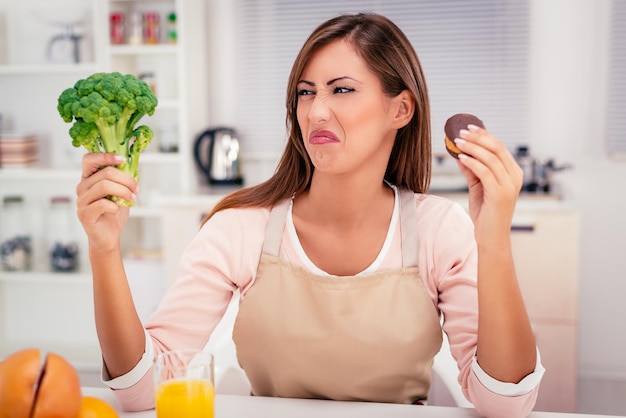 Mooie jonge vrouw in de keukenbeslissing tussen broccoli of zoete chocoladetaart.