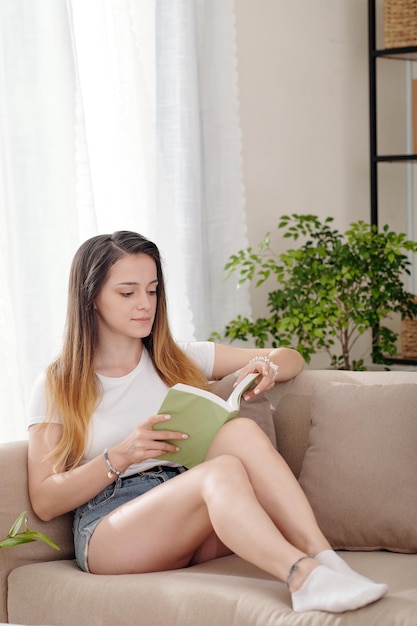 Mooie jonge vrouw in denim shorts zittend op de bank en het lezen van een boek met verhalen