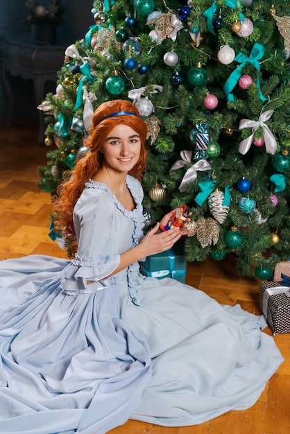 mooie jonge vrouw in een baljurk zit bij de kerstboom met een speelgoed en glimlacht