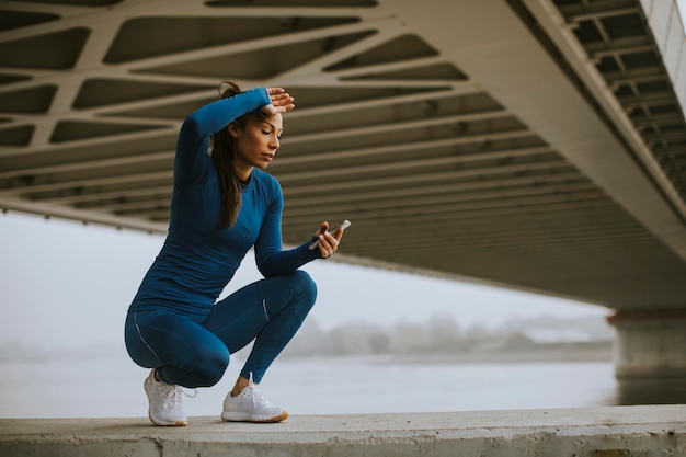 Mooie jonge vrouw in een blauw trainingspak die haar trainingsvoortgang volgt op de fitness-app bij de rivier in de herfstochtend