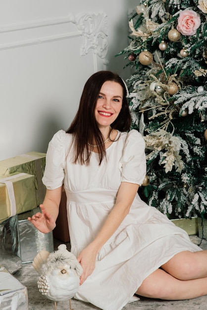 Mooie jonge vrouw in een elegante jurk die naast de kerstboom en cadeautjes staat.