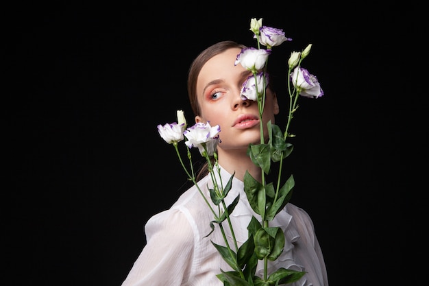 Mooie jonge vrouw in een elegante witte blouse die haar gezicht aanraakt met natuurlijke bloemen en wegkijkt tegen een zwarte achtergrond