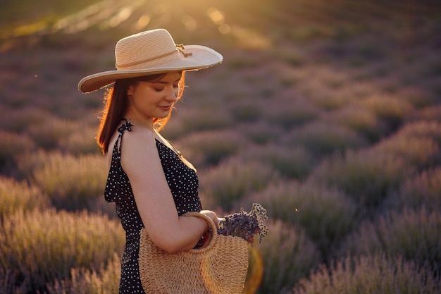 Mooie jonge vrouw in een hoed en tas met lavendelbloemen op een veld tijdens zonsondergang