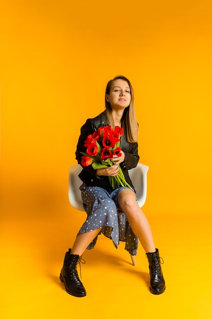 Mooie jonge vrouw in een jurk en een zwart leren jasje zit op een stoel met rode tulpen op een gele muur