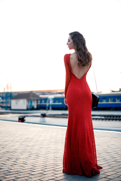 Mooie jonge vrouw in een lange rode jurk staat op het perron van het station bij de spoorweg