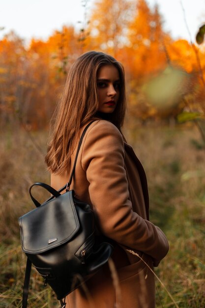 Mooie jonge vrouw in een modieuze herfstbeige jas met een leren rugzak reist in de natuur met herfstgebladerte