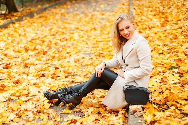 Mooie jonge vrouw in een trendy klassieke jas in stijlvolle schoenen zit in de buurt van een gele herfstbladeren