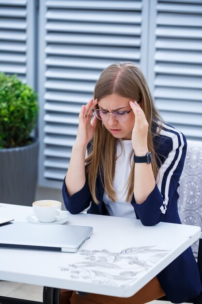 Mooie jonge vrouw in een wit T-shirt drinkt koffie, ze heeft hoofdpijn en werkt op een laptop en zit buiten in een café. Jonge vrouwenlaptop voor het werk. Zakelijke problemen