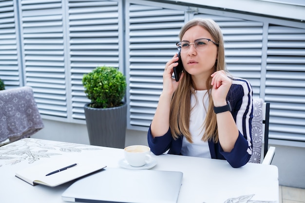 Mooie jonge vrouw in een wit T-shirt drinkt koffie, ze heeft hoofdpijn en werkt op een laptop en zit buiten in een café. Jonge vrouwenlaptop voor het werk. Zakelijke problemen