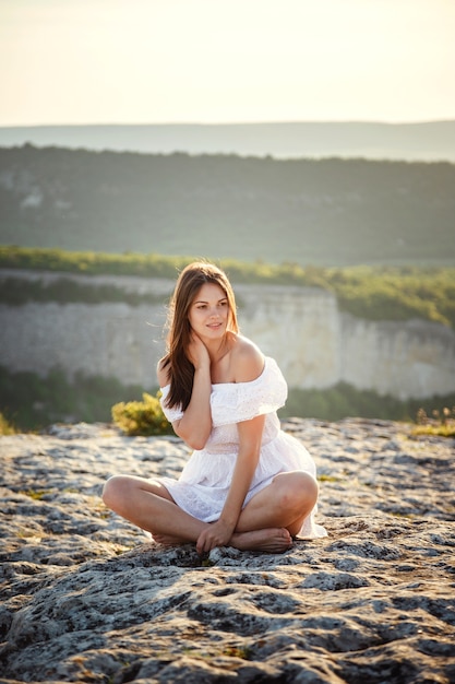 Mooie jonge vrouw in een witte jurk geniet van een prachtig landschap in de bergen tijdens zonsondergang