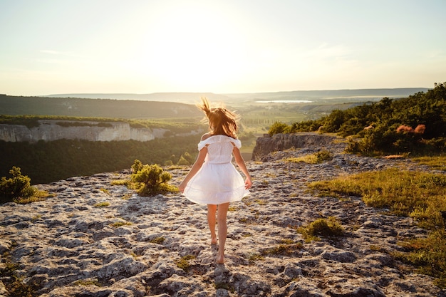 Mooie jonge vrouw in een witte jurk geniet van een prachtig landschap in de bergen tijdens zonsondergang