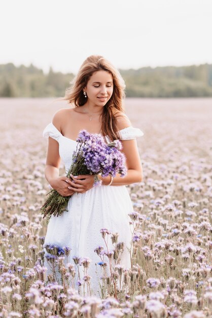 Mooie jonge vrouw in een witte jurk met een boeket bloemen in een veld in de zomer in de natuur