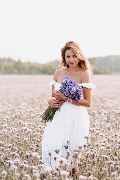 Mooie jonge vrouw in een witte jurk met een boeket bloemen in een veld in de zomer in de natuur
