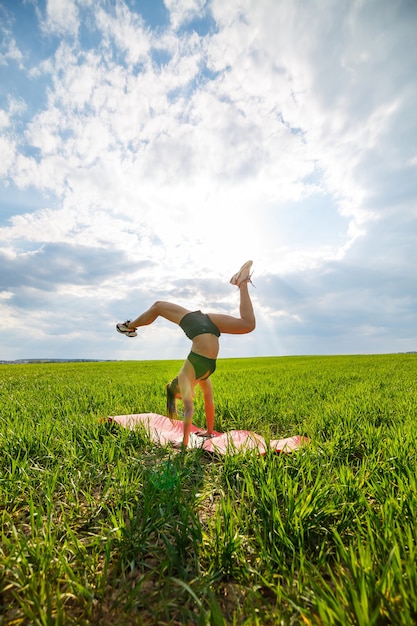 Mooie jonge vrouw in een zwarte top en korte broek voert een handstand uit. Een model staat op haar handen en doet gymnastische splitsingen tegen de blauwe lucht. Gezond levensstijlconcept