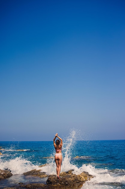 Mooie jonge vrouw in een zwembroek op een rotsachtig strand op een zonnige dag tegen de achtergrond van golven Vakantie in het zomerseizoen Selectieve aandacht