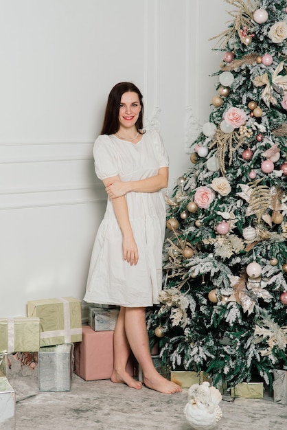 Mooie jonge vrouw in elegante jurk staande naast kerstboom en cadeautjes.