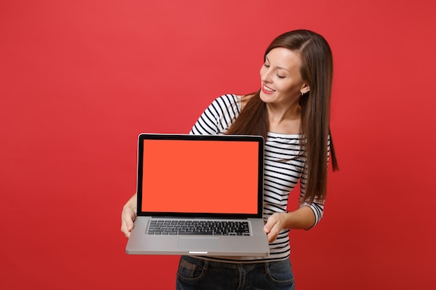 Mooie jonge vrouw in gestreepte kleding op zoek op laptop pc-computer met leeg zwart leeg scherm in handen geïsoleerd op rode achtergrond. Mensen oprechte emoties, lifestyle concept. Bespotten kopie ruimte.