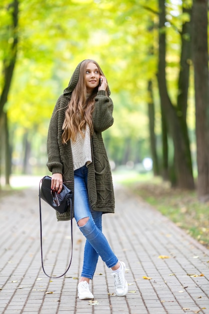 Mooie jonge vrouw in groen vest met een kap die in de herfstpark loopt en mobiele telefoon spreekt.