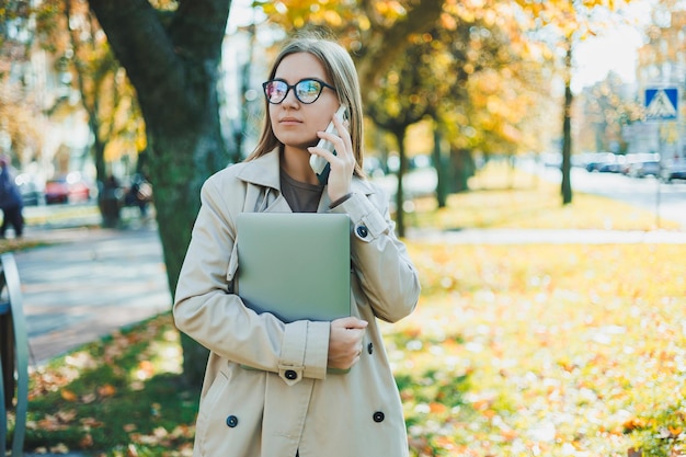 Mooie jonge vrouw in herfstjas die laptop en telefoon gebruikt die op afstand werkt vrouw praten op mobiele telefoon in herfstpark