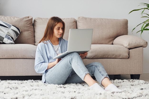 Mooie jonge vrouw in jeans en blauw shirt zit op de vloer met laptop binnenshuis thuis