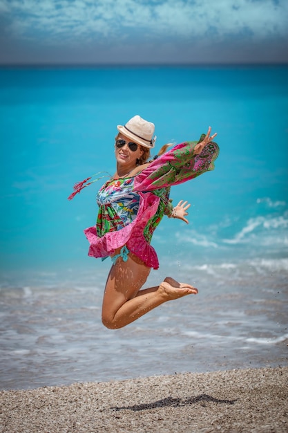 Mooie jonge vrouw in kleurrijke jurk speelt in de kust op het strand tijdens de hete zomervakantiedag. Ze geniet en springt op het strand.