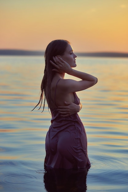 Mooie jonge vrouw in meerwater in zomerjurk bij zonsondergang Portret van een romantisch nat meisje bij zonsondergang warme zon natuurlijke schoonheid van een vrouw