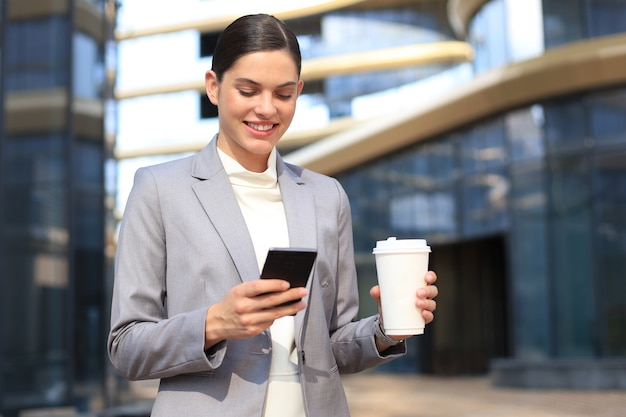 Mooie jonge vrouw in pak met behulp van slimme telefoon en glimlachen terwijl je buiten staat.