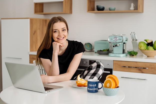 Mooie jonge vrouw in pyjama die 's ochtends met laptop in de keuken werkt