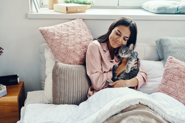 Mooie jonge vrouw in pyjama glimlachend en omhelzend huiskat terwijl ze thuis in bed rust