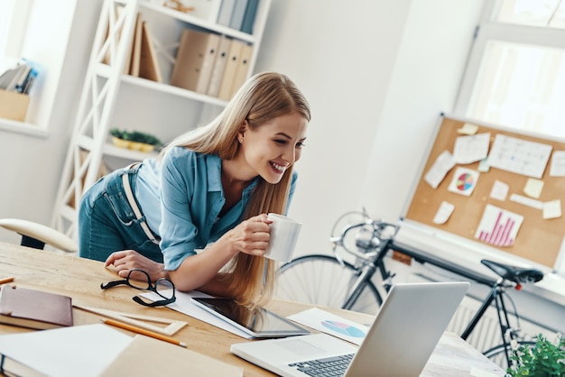 Mooie jonge vrouw in slimme vrijetijdskleding die lacht en laptop gebruikt terwijl ze op kantoor werkt